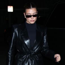 Hailey Bieber wearing a black Khaite leather trench coat with a turtleneck, sunglasses, and black Saint Laurent boots and clasp bag.