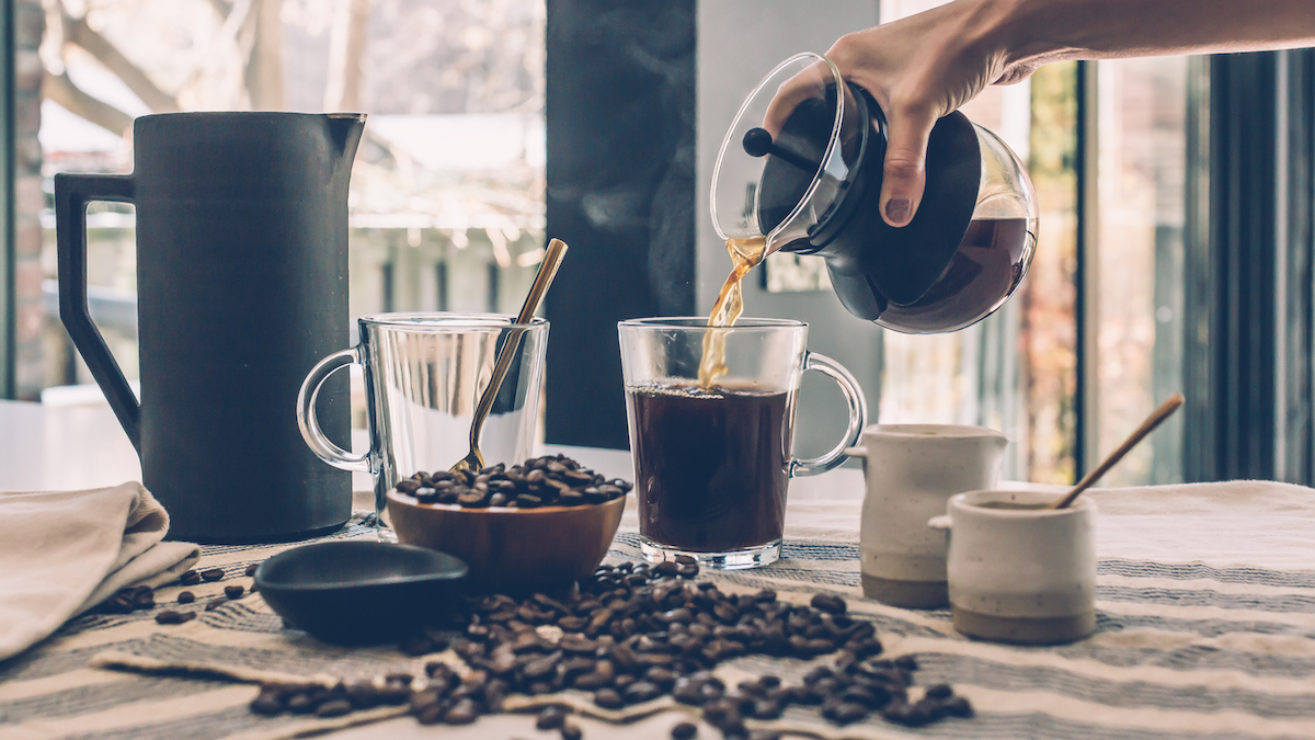 Seseorang menuangkan kopi ke dalam cangkir kaca yang dikelilingi oleh biji kopi