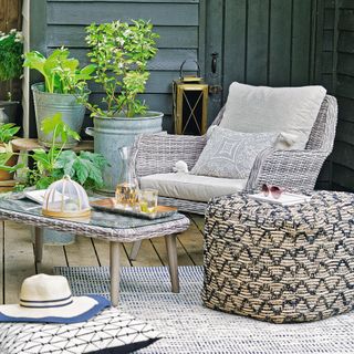 A sheltered outdoor seating area on a decking terrace with a wicker chair, coffee table and ottoman with straw hat on a cushion with zigzag patterns