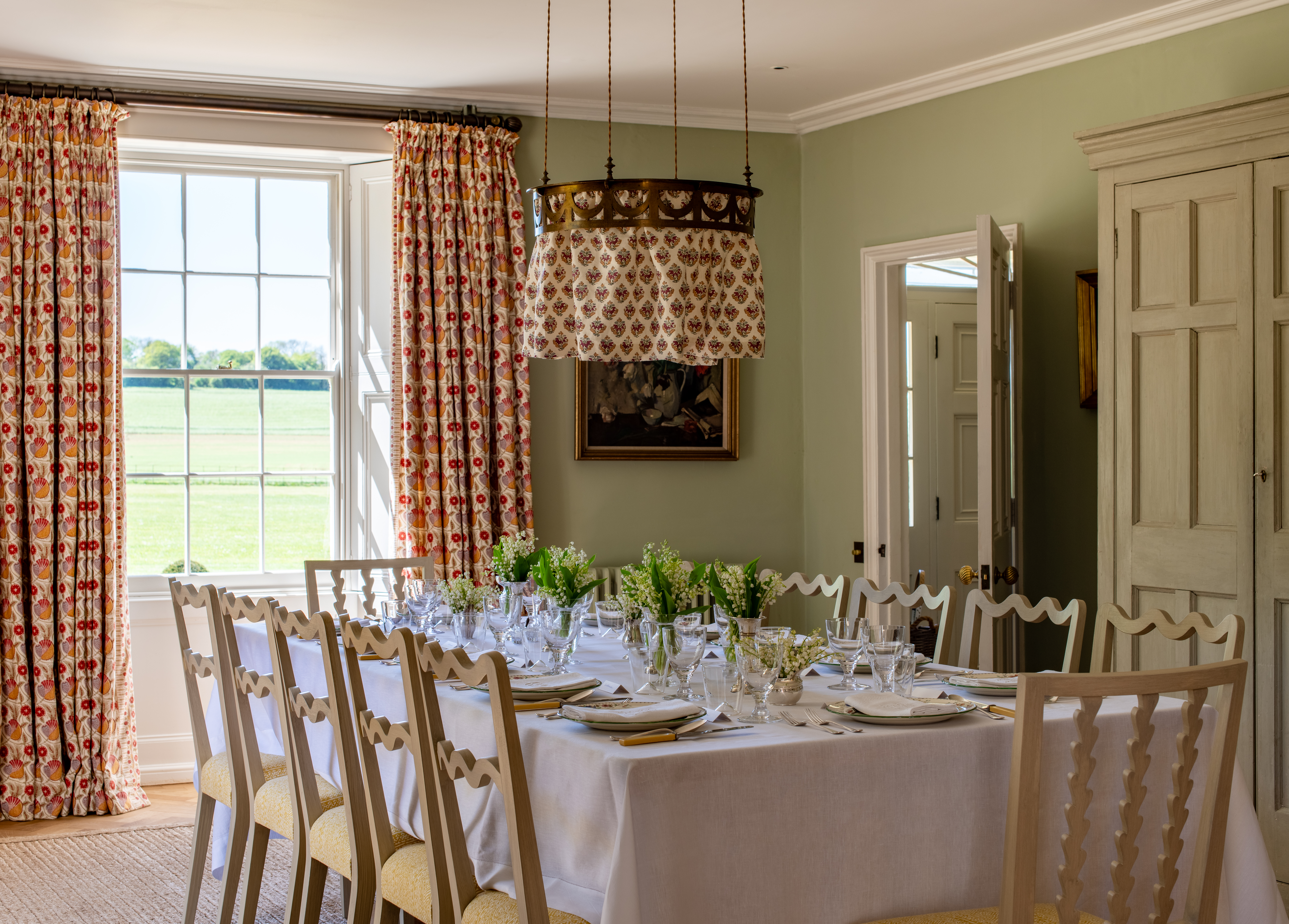 A dining rooms with a table, covered in a white cloth, wavy back chairs, a cloth pendant light and matching curtains