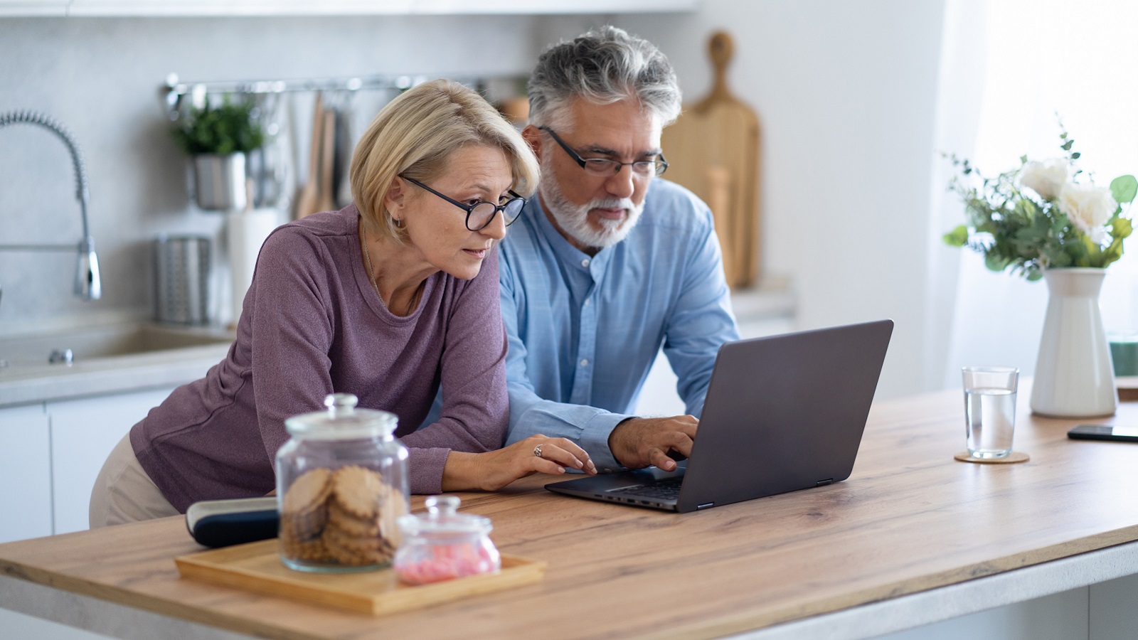 An older couple in the kitchen look at a laptop computer.
