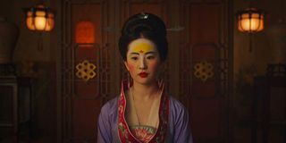 Mulan, wearing traditional Chinese makeup, stands in a dimly lit room in a scene from the live actio