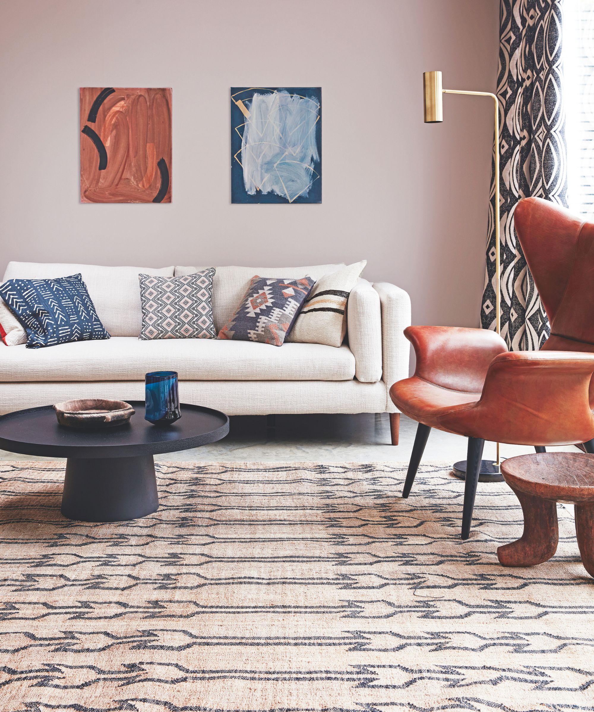 A living room with patterned rug, black circular coffee table with a bowl and blue vase on top, a white couch with patterned cushions, brown leather chair and wooden table next to it. To the back right is a window with patterned windows, and a gold floor lamp. Above the sofa are two abstract artworks.