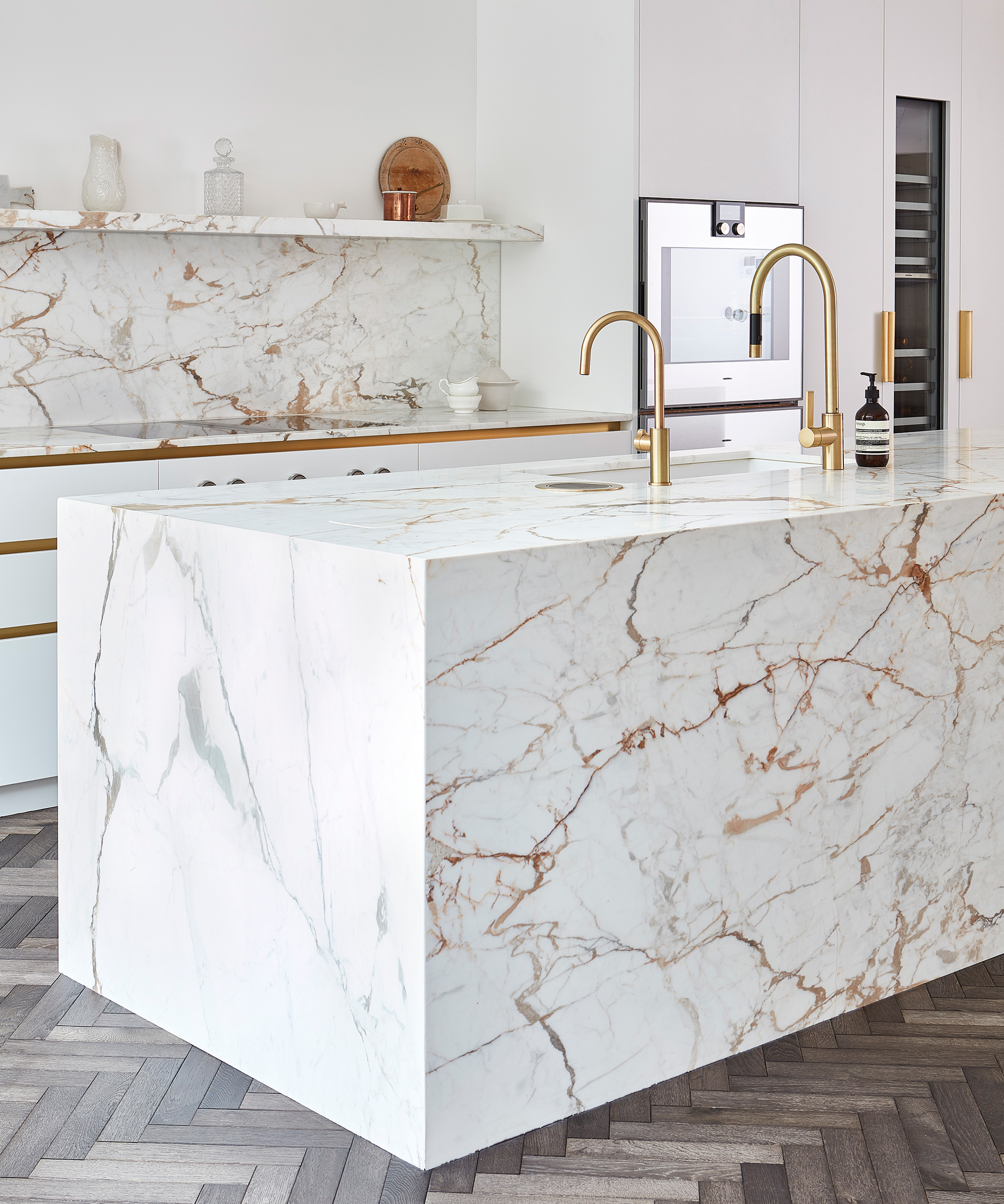 A marble island and backsplash with gold faucets and herringbone flooring