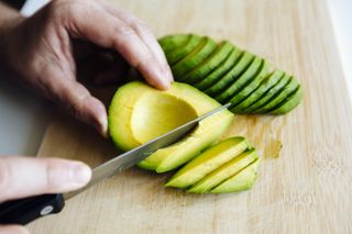 A close up shot of a man's hands slicing an avocado on a board with a knife.