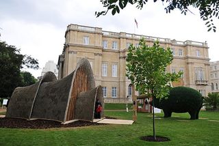 The garden area of London's Marlborough House featuring a double igloo style house made with shells and branches, a grass brush trimmed in the shape of a pig, and the building in the backdrop.