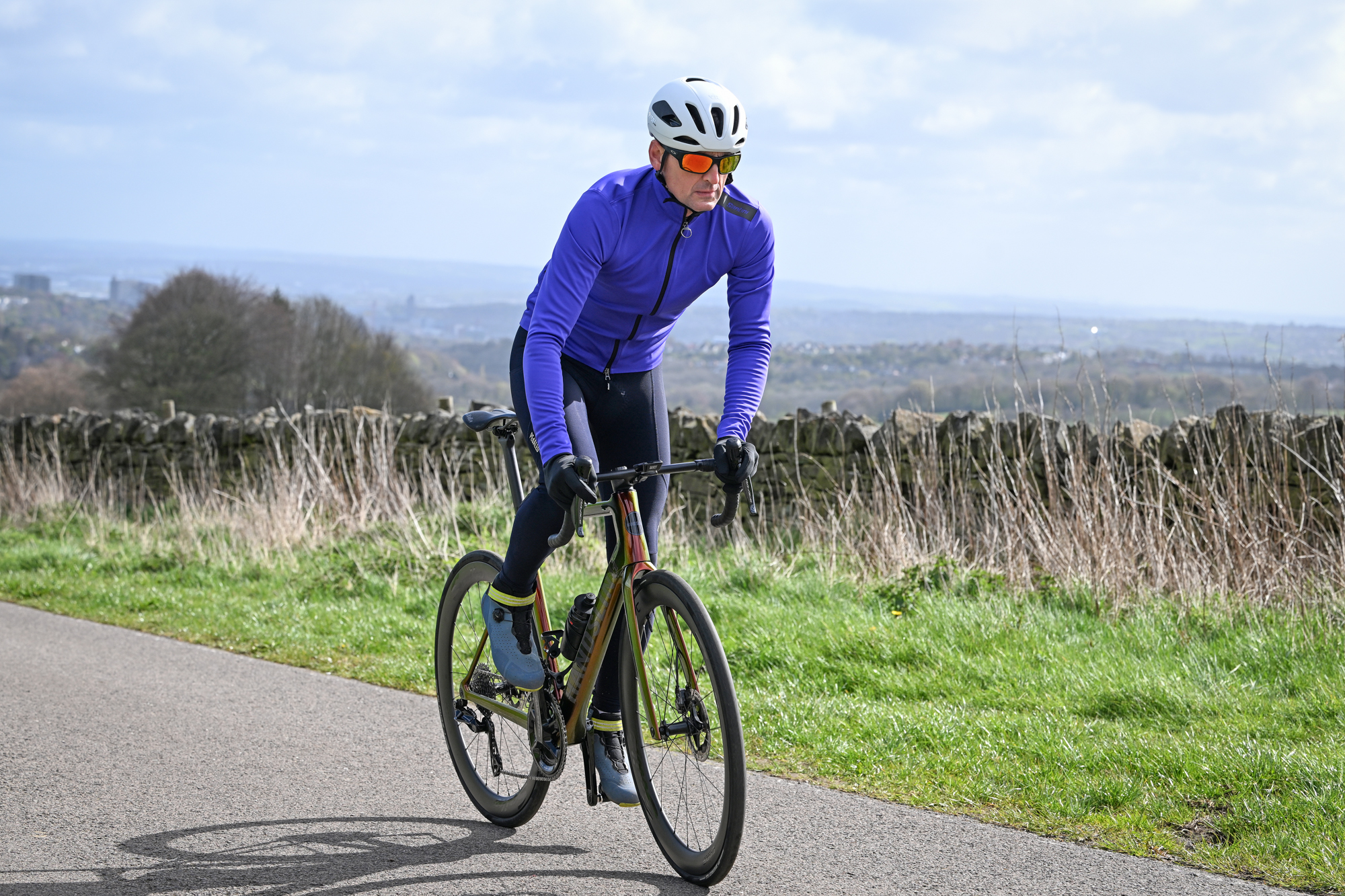 Man in a purple jacket, sunglasses and white helmet riding a bike up a country lane out of the saddle