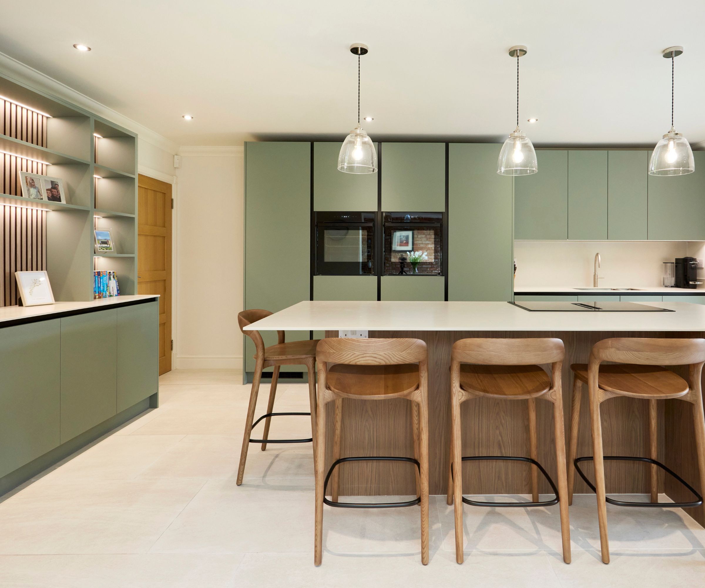 A spacious open-plan kitchen with sage green handleless cabinetry, a large white-topped island with oak bar stools, glass pendant lights and large format pale stone floor tiles.