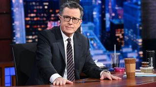 Stephen Colbert sits at his desk in a suit and stares at the camera during the finale Late Show week. 