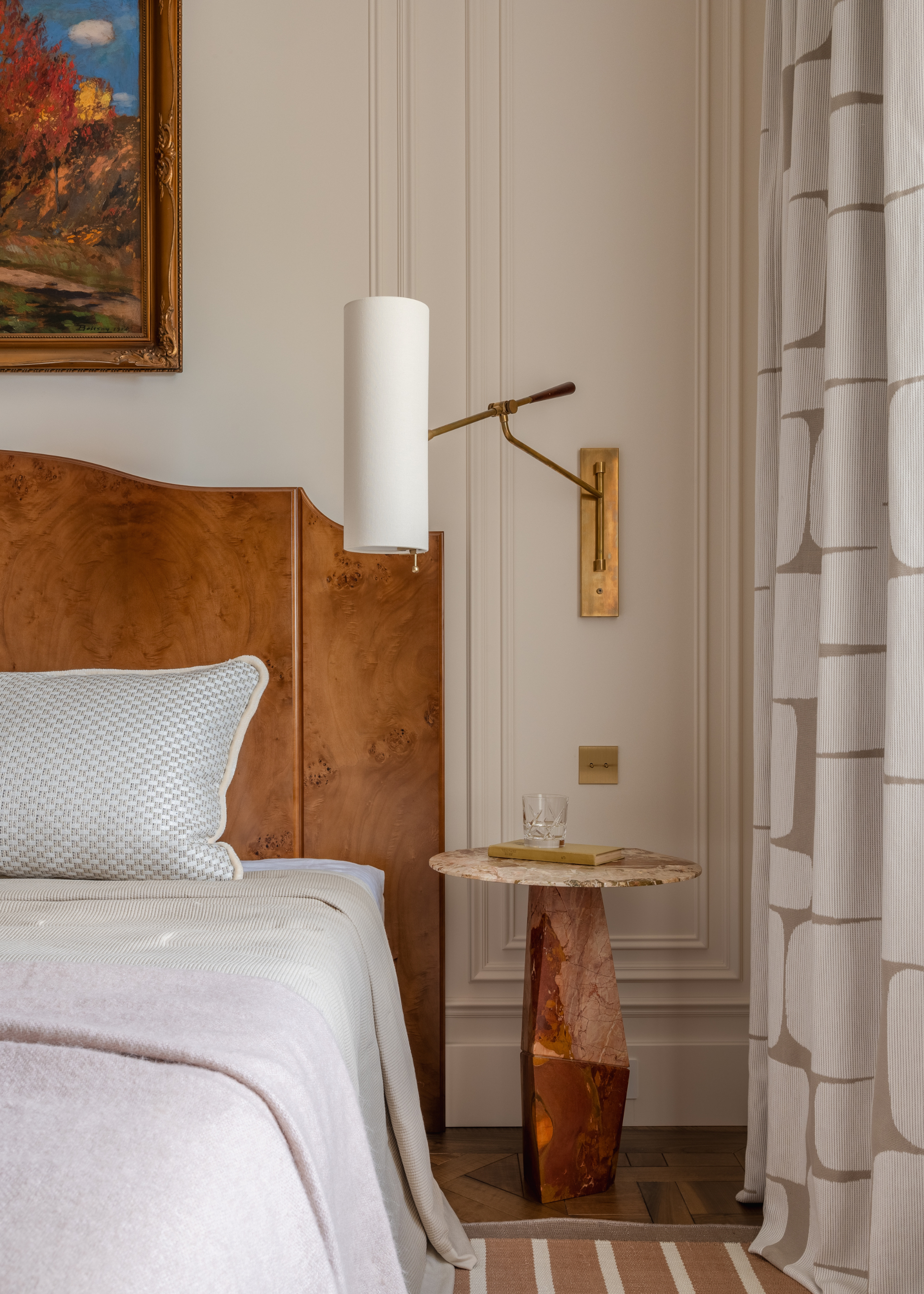 An off-white bedroom with a wood headboard, a stone side table, an extendable wall sconce, a striped rug, patterned curtains, and a framed painting