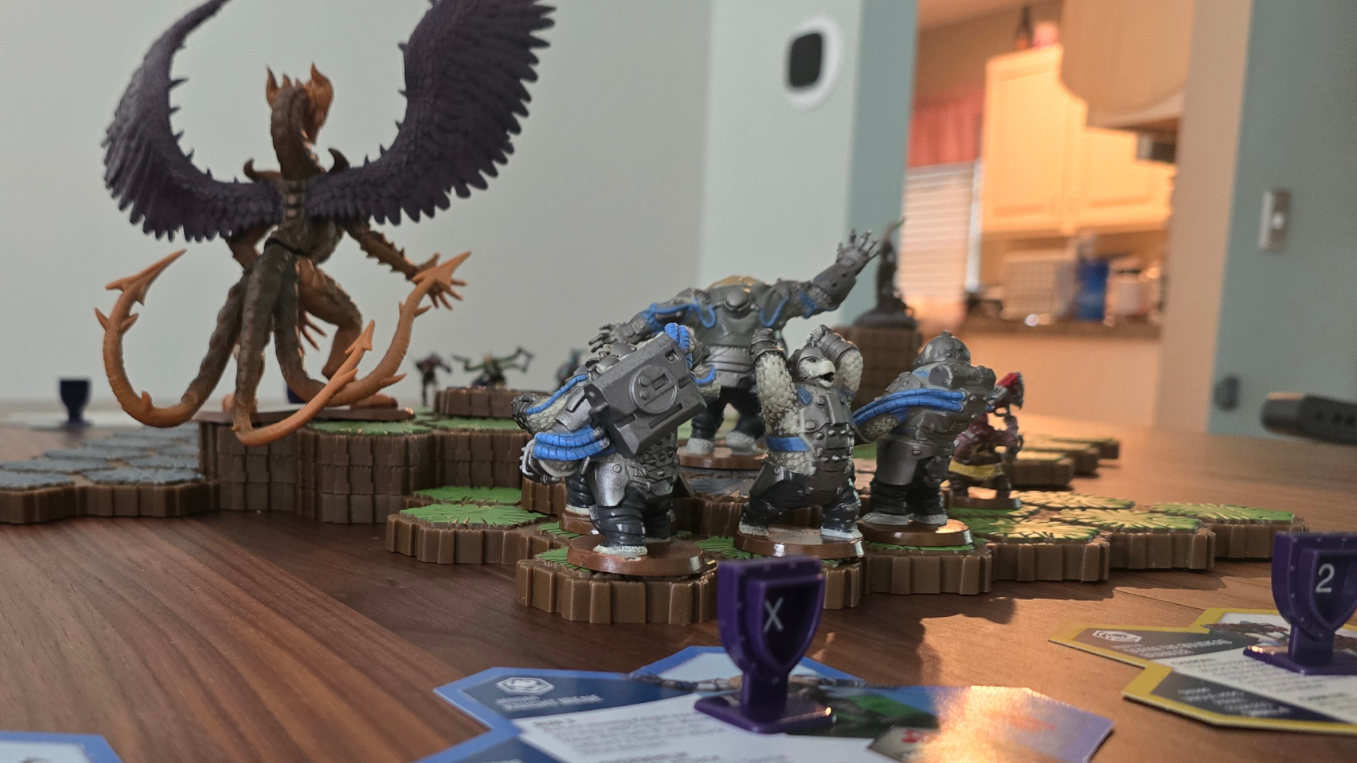 A selection of Heroscape miniatures, terrain, tokens, and components on a wooden table, with a kitchen visible in the background
