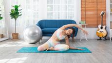 woman in a tie-dyed crop top and leggings performing a pilates mermaid stretch on a blue mat in a living room setting. 