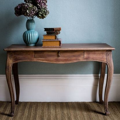 Marie Console Table in mango wood with long thin drawer, set against a blue wall and with a vase of flowers and books on top