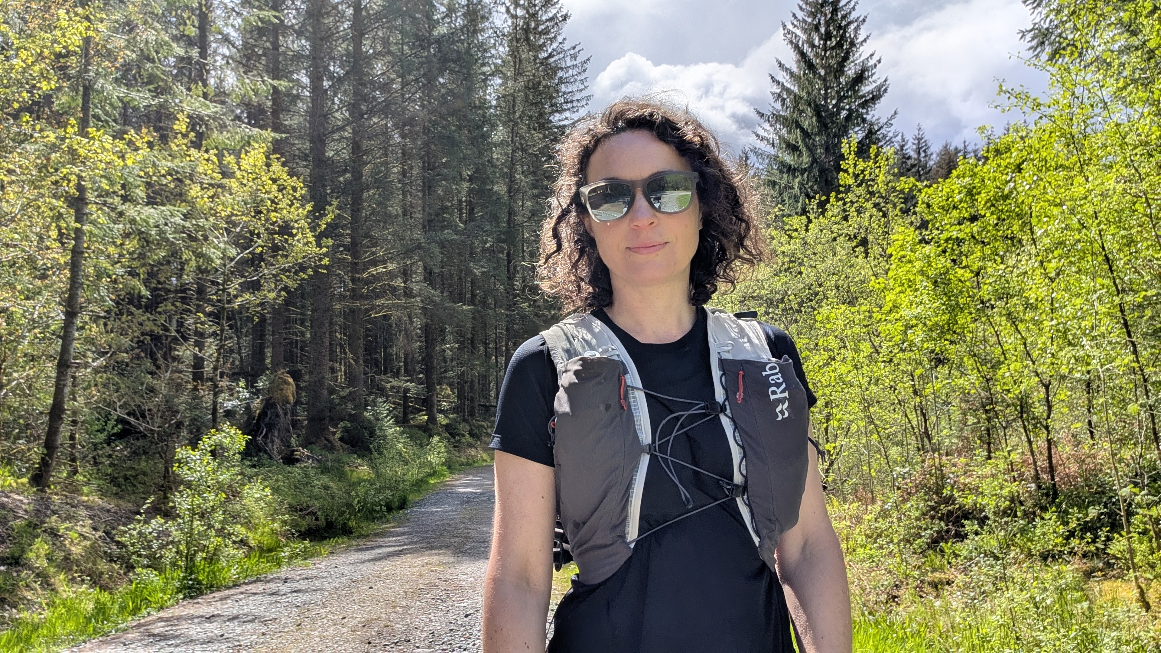 A woman trail runner wearing a black T-shirt and a grey Rab running vest on a forest trail