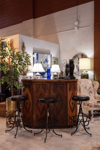 An indoor flea market features high-end furniture and antiques, including a refined wooden bar, wrought iron and leather stools, and Art Deco lamps.