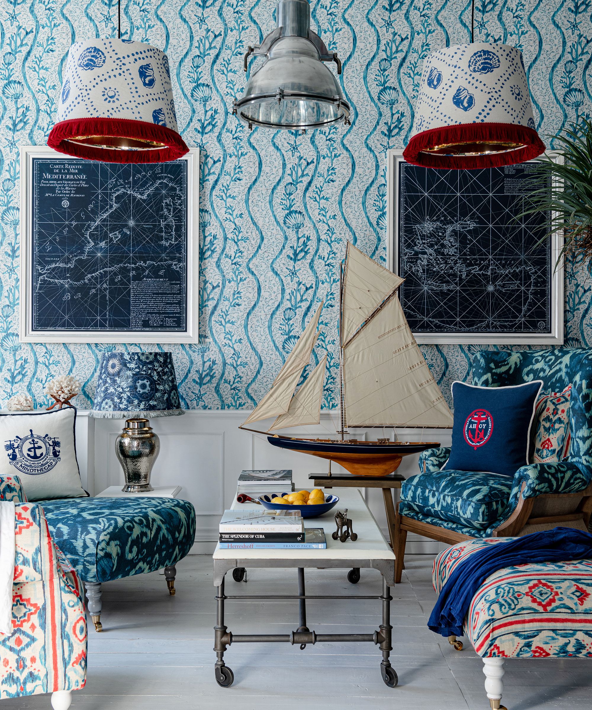 A small sitting room decorated in blue and white, with patterned wallpaper showing shells and waves. Two upholstered chairs face a low table. Model sailing boats, framed maps and fabric lampshades create a seaside, nautical atmosphere with traditional coastal character.