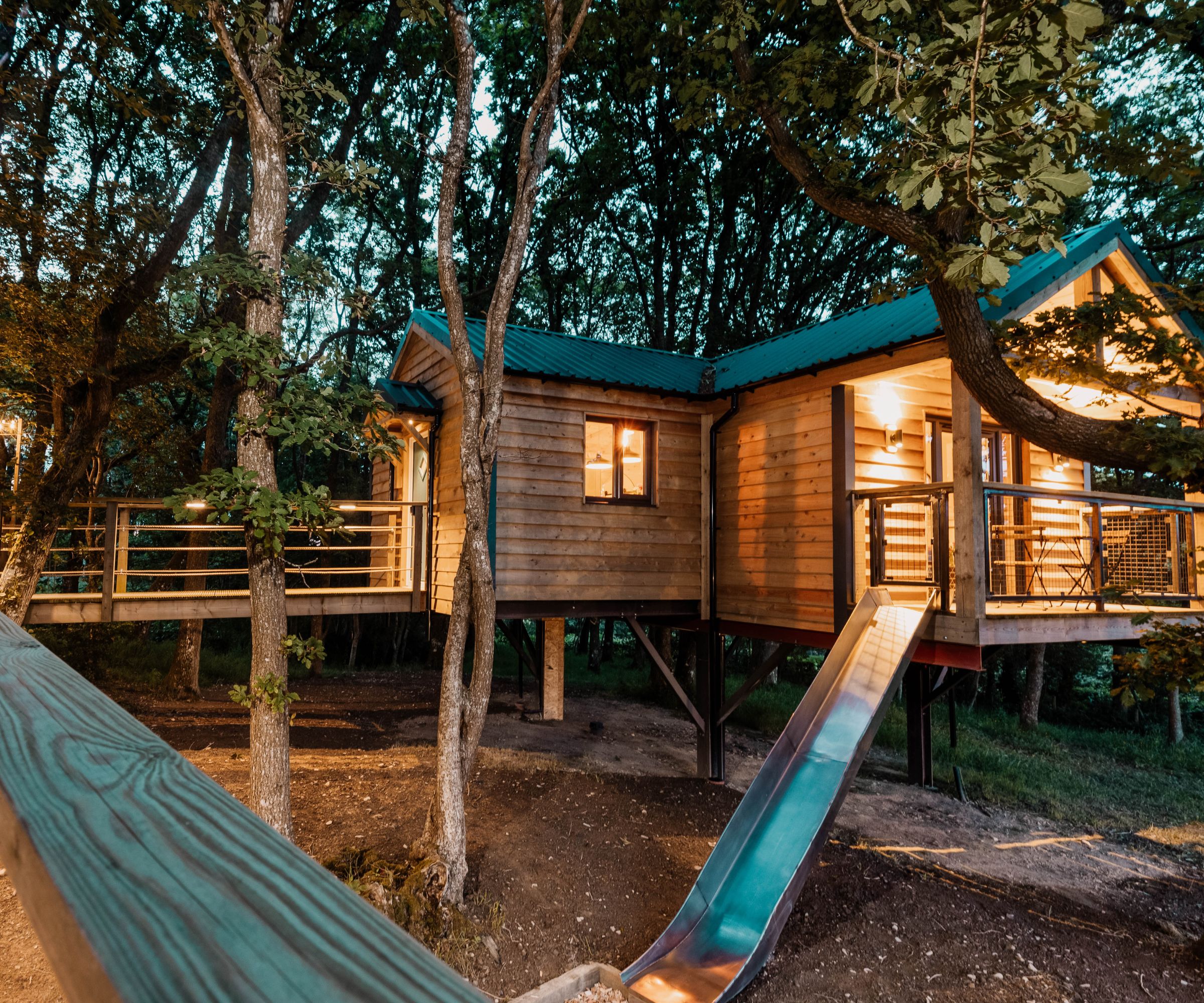 A large wooden treehouse with multiple sections, a green roof, a wraparound deck, and a metal slide, set among tall trees.