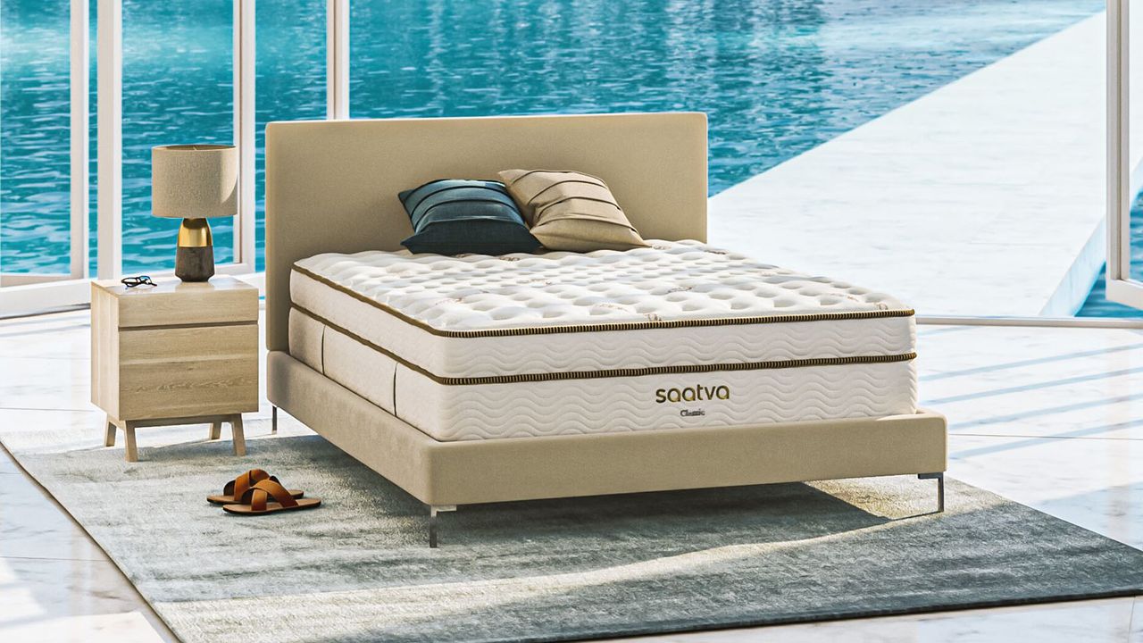 Image shows the Saatva Classic on a beige fabric bed frame in a sunny outdoor room overlooking a blue swimming pool on a bright day