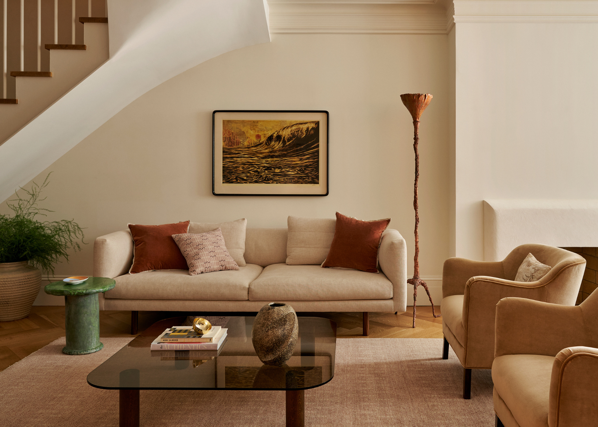 A white living room with cream furniture, a low-glass table, a vase, a stack of books, a green side table, a potted plant, a framed painting, a standing lamp and a pair of armchairs on a beige area rug