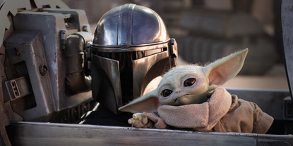 The Mandalorian in full armour including a helmet, with Grogu, a green alien with elf-like ears sitting beside him.