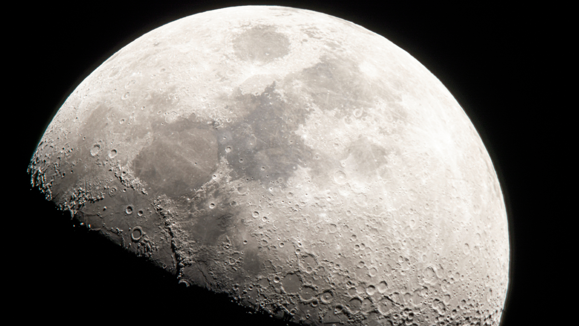 A close up of the moon in the darkness of space, its craters and dark maria visible against the gray surface, while its bottom half is covered in shadow.