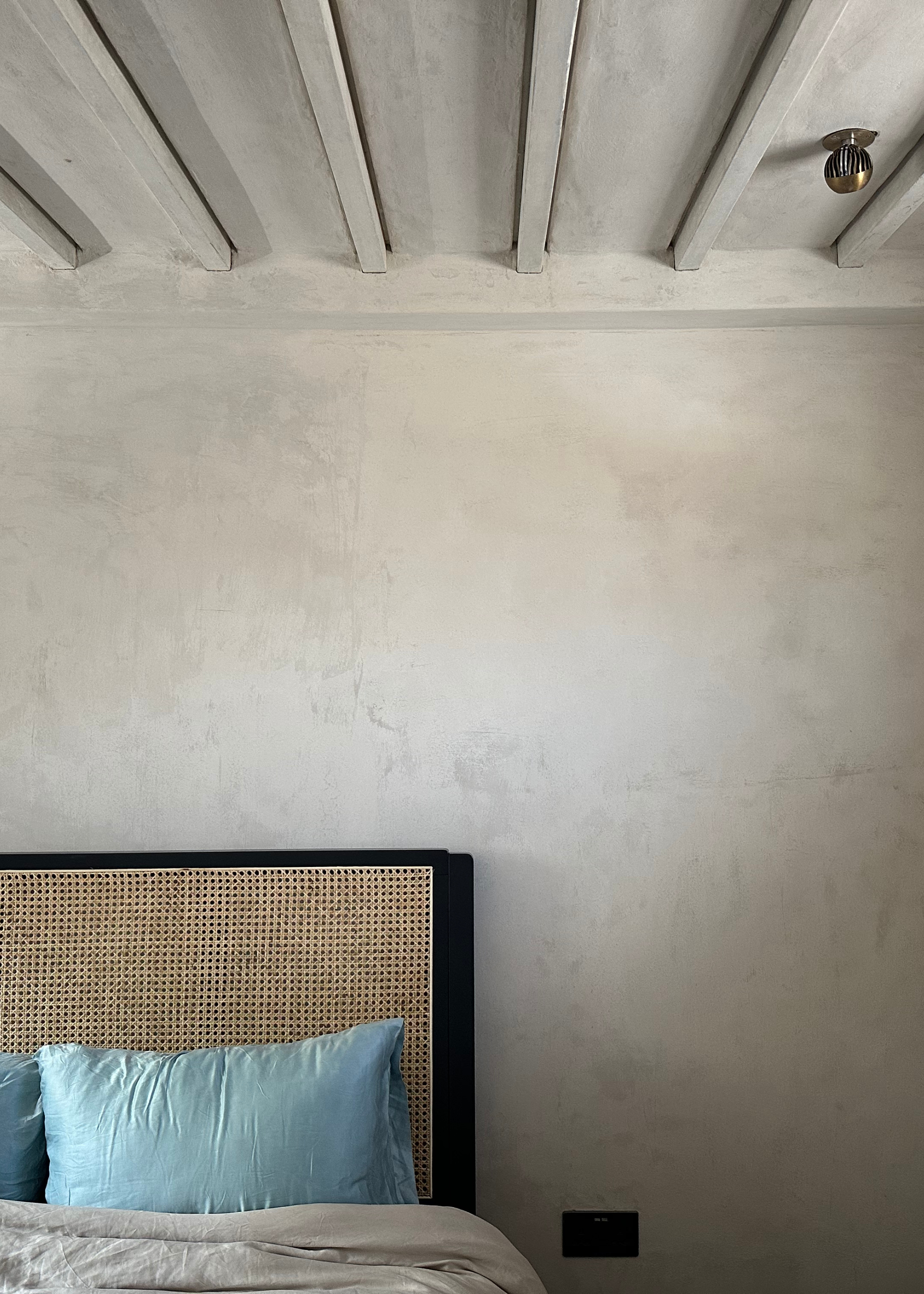 A bedroom with a rattan headboard and sky blue pillows, exposed ceiling beams and brutalist cement walls