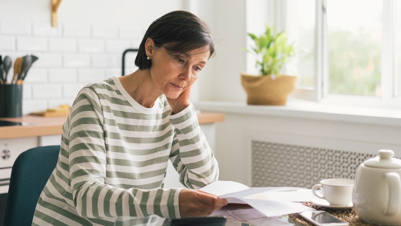 An older woman looks over paperwork at the kitchen table with a pensive look on her face.