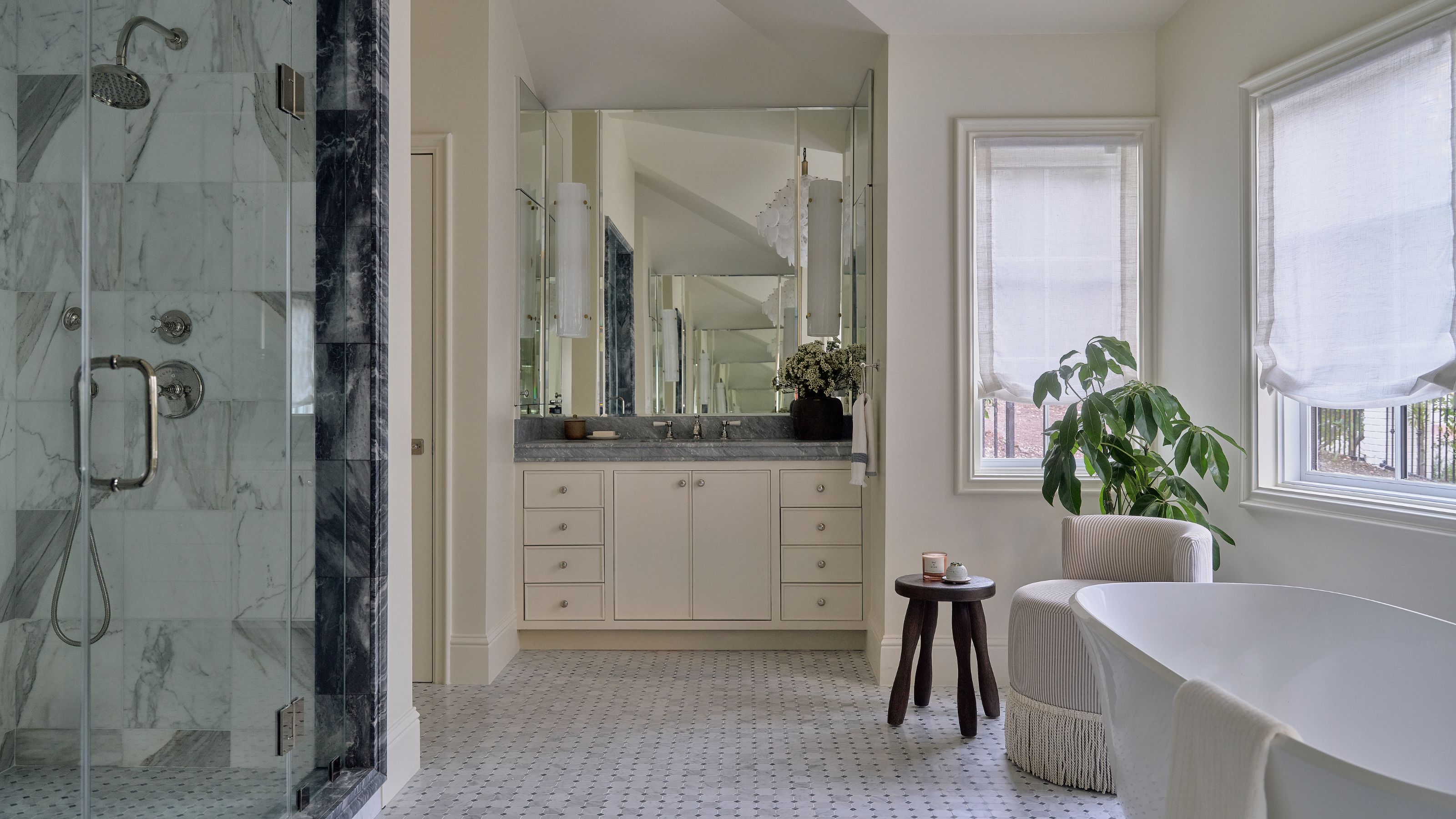 a large white primary suite bathroom with a marble shower, freestanding tub, vanity unit and a small fringed chair with a wooden stool