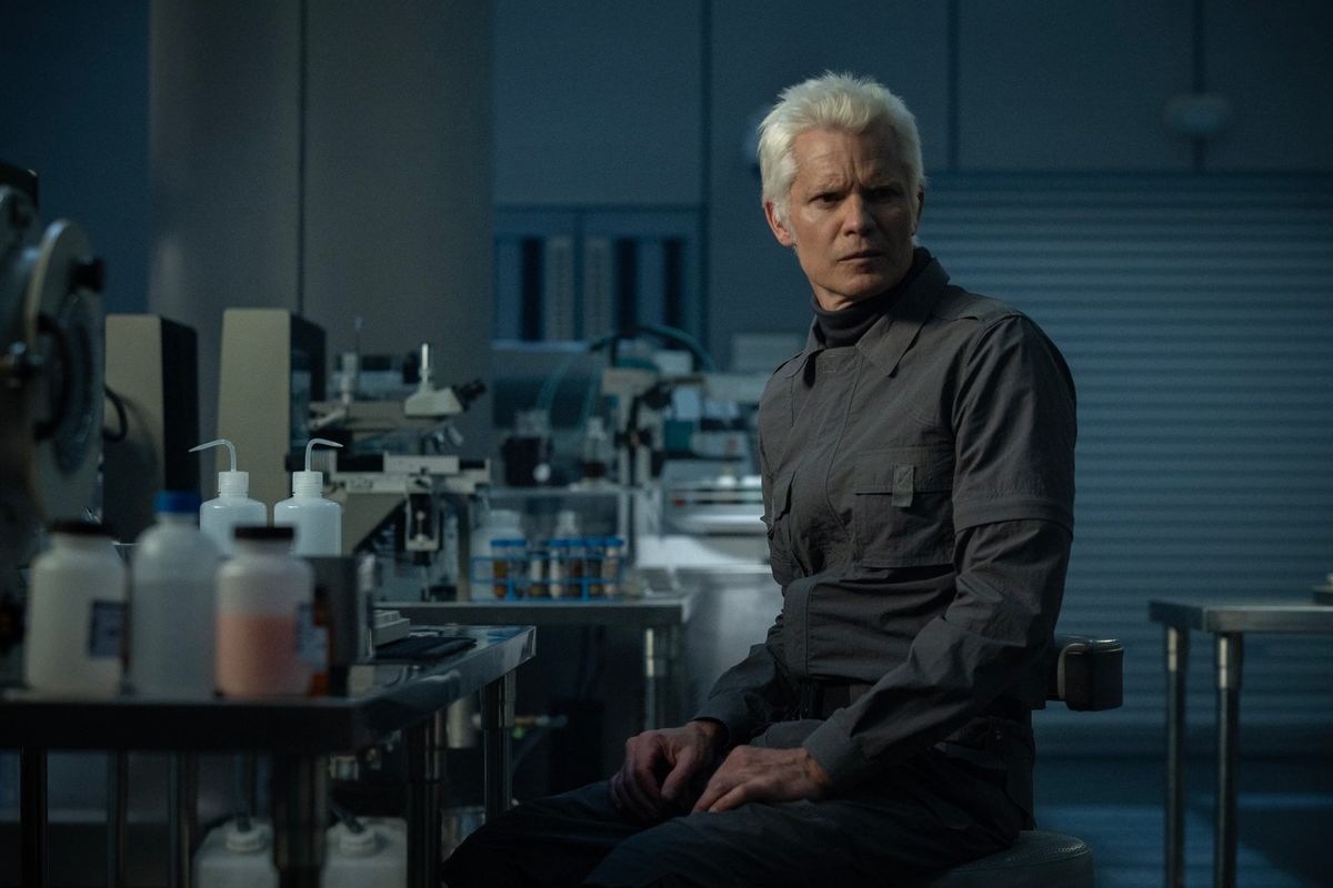 Still from the TV show Alien: Earth. An older man is sitting on a stool in a lab. He is wearing a gray jumpsuit and has short white hair.