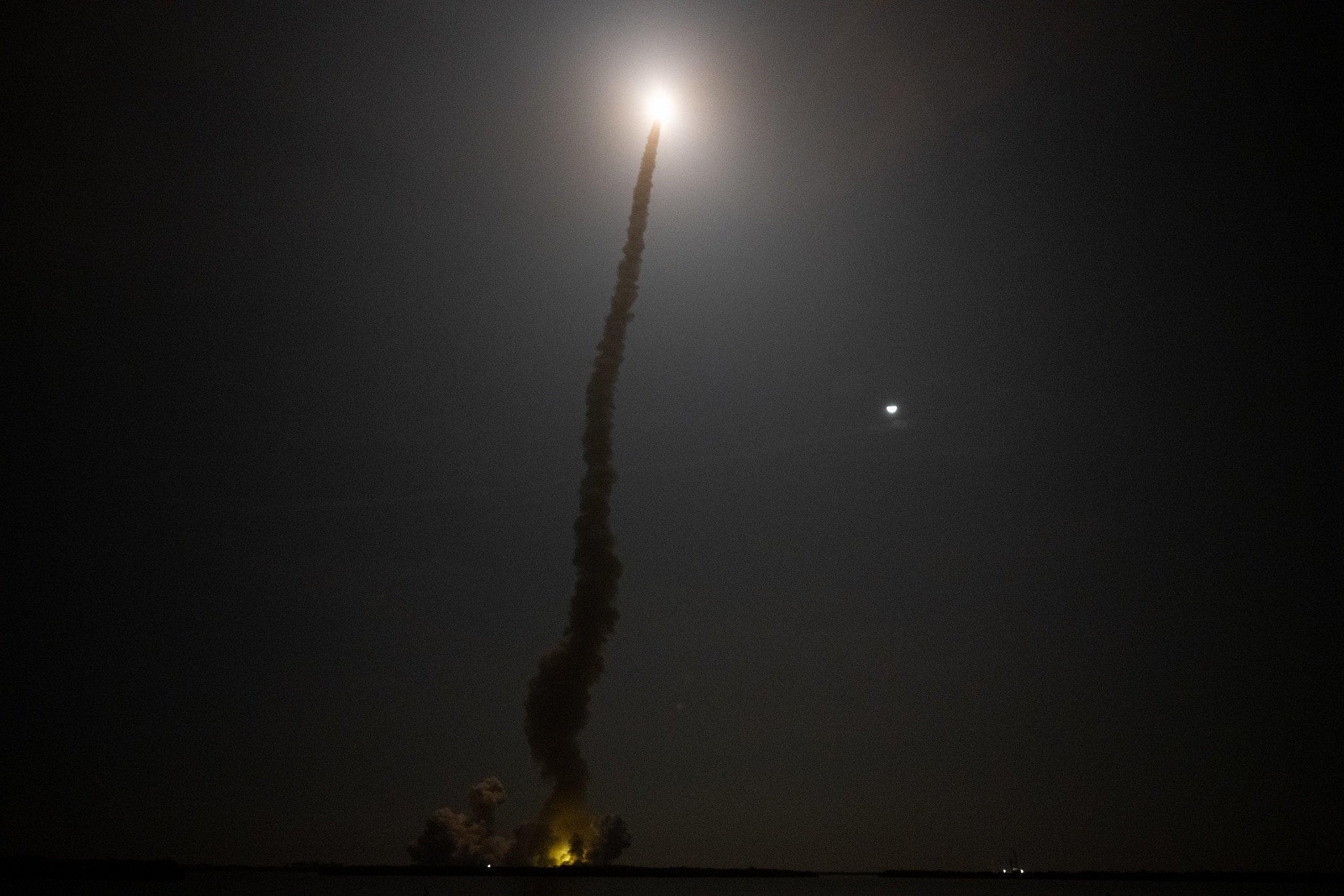 NASA's SLS rocket heads towards deep space as the moon shines beside its smoke trail in the night sky.