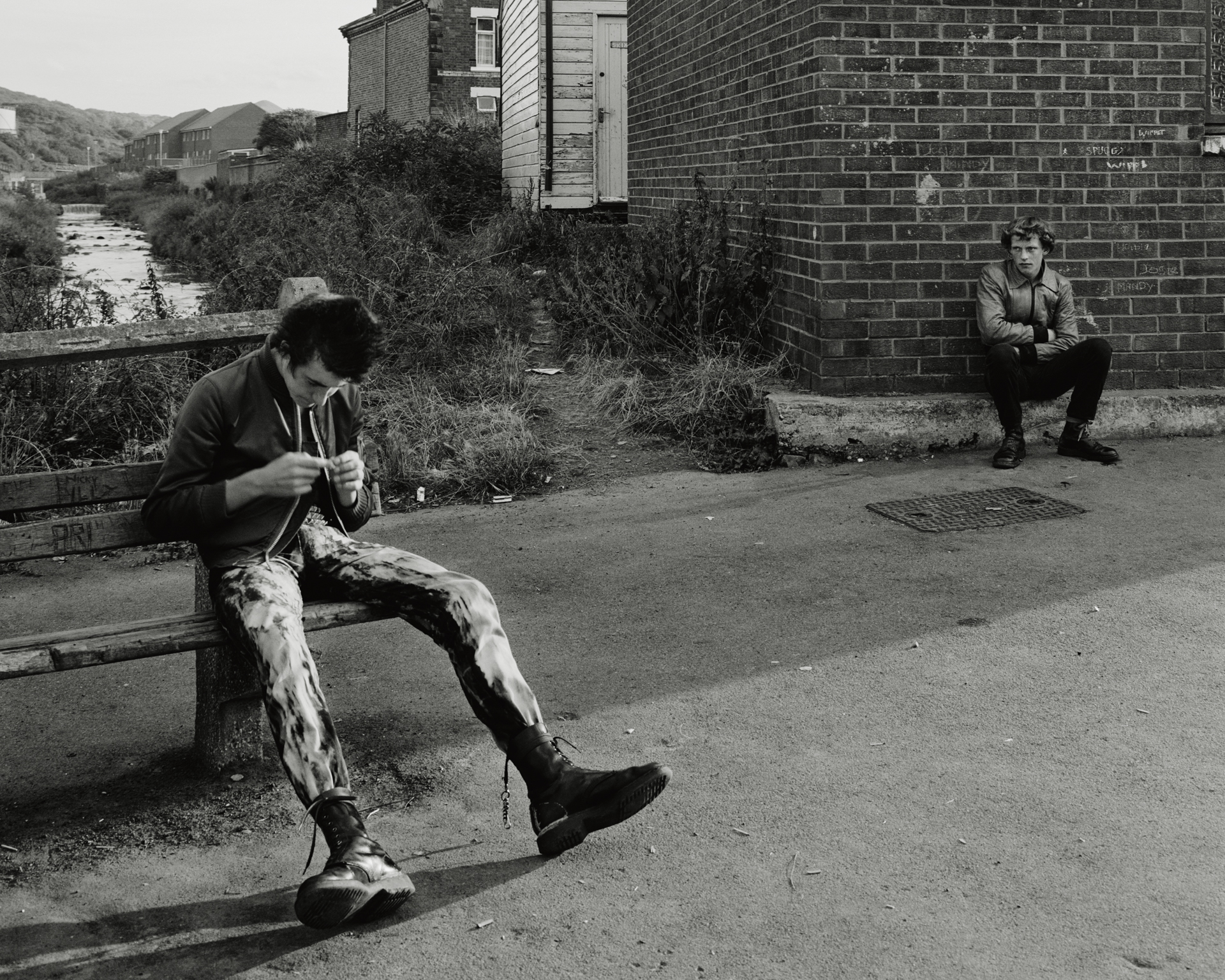 Black and white photograph of a young man in patterned trousers sitting on a bench beside a stream, absorbed in something in his hands, while another young man watches from a low wall in the background, taken at Skinningrove, North Yorkshire, in 1982.