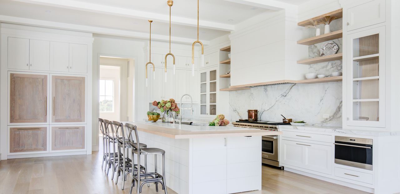 A luxury spacious white kitchen with an island, a marble splashback and light wooden finishes 