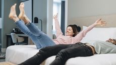 A man in a green shirt lies on a mattress as a woman in a pink top jumps happily onto the bed next to him