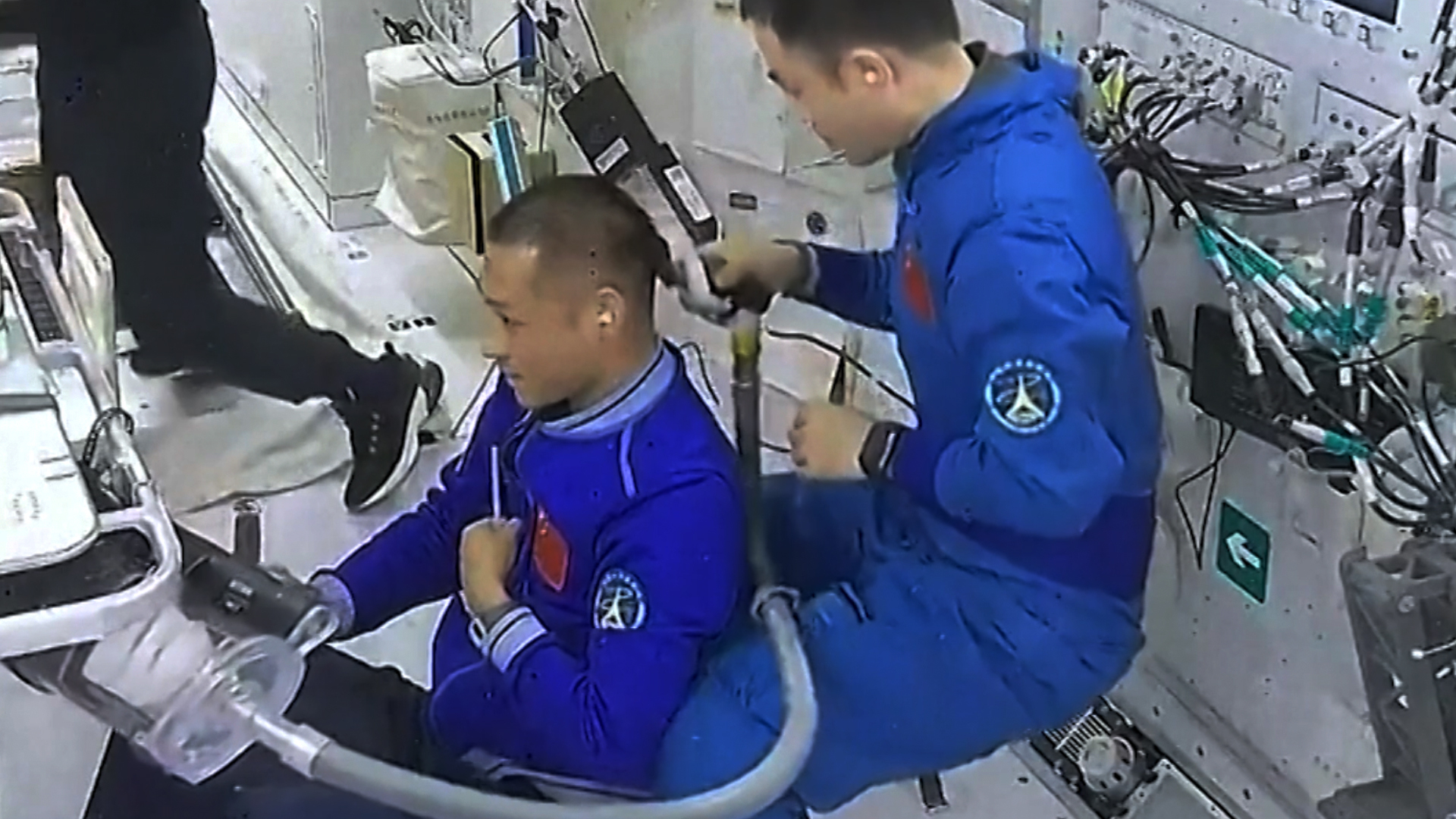 un astronauta chino con un traje de vuelo azul le corta el pelo a su colega vestido de manera similar dentro de una estación espacial.