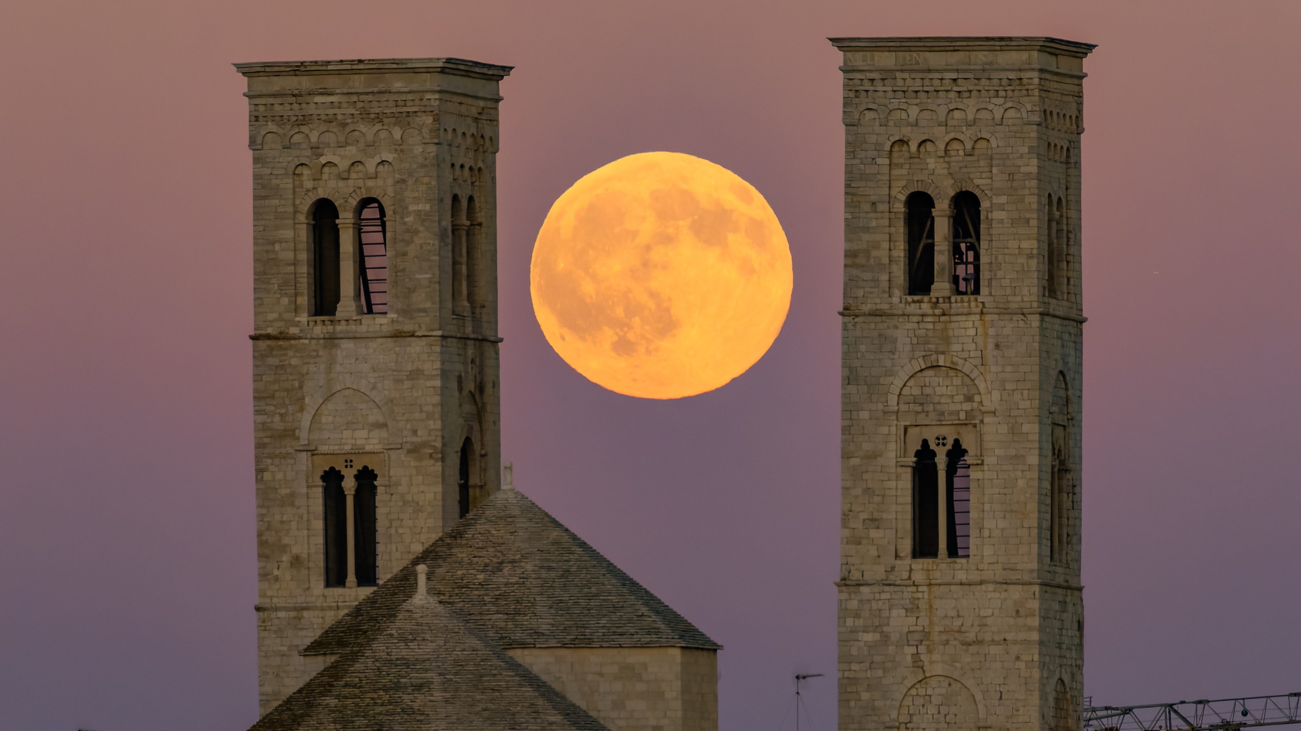 A large orange full moon rises between the two towers of Molfetta Cathedral against a dark blue and pink sky in Molfetta, Italy.