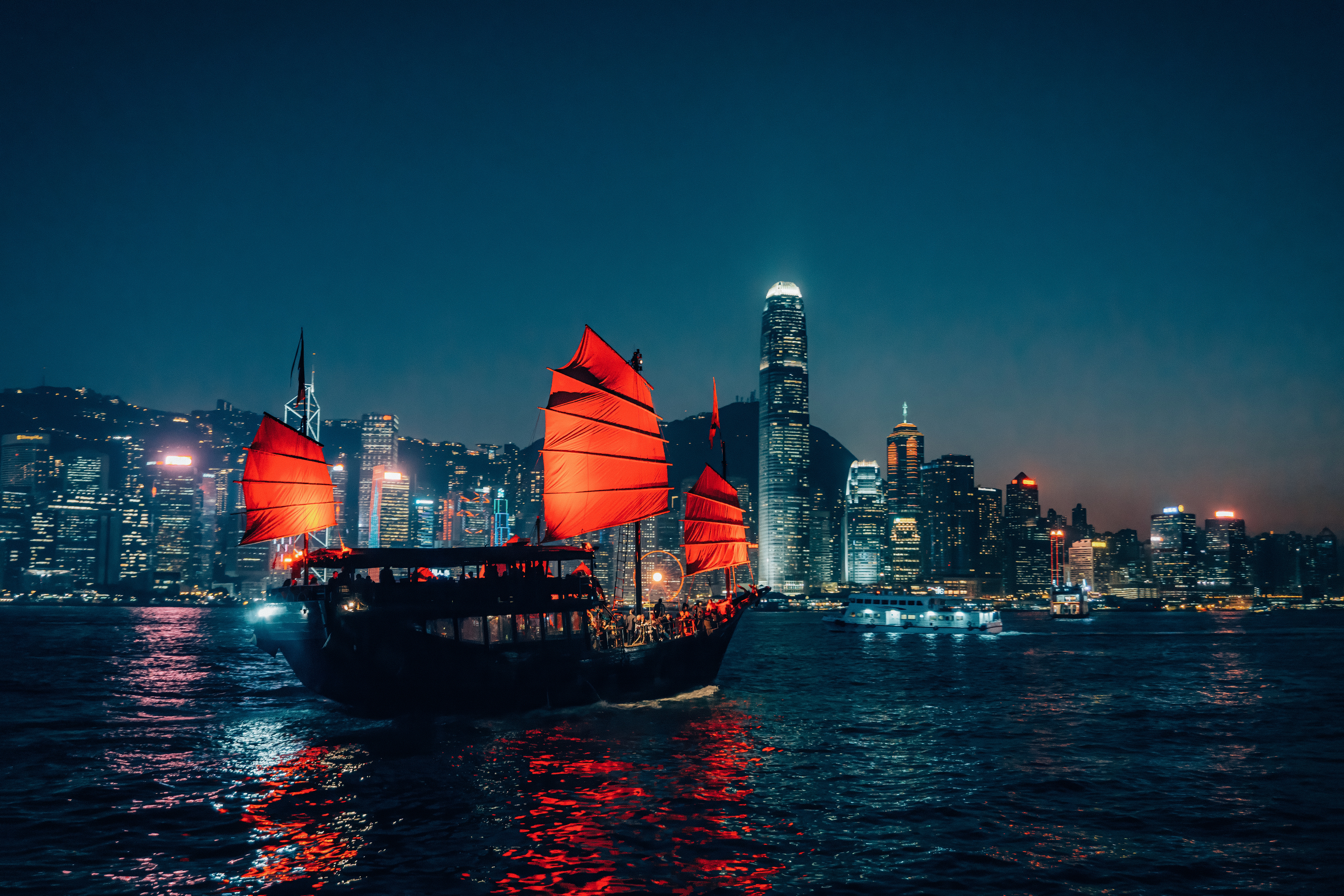 A vintage boat with red sails sails into the sea at night against beautifully lit skyscrapers.