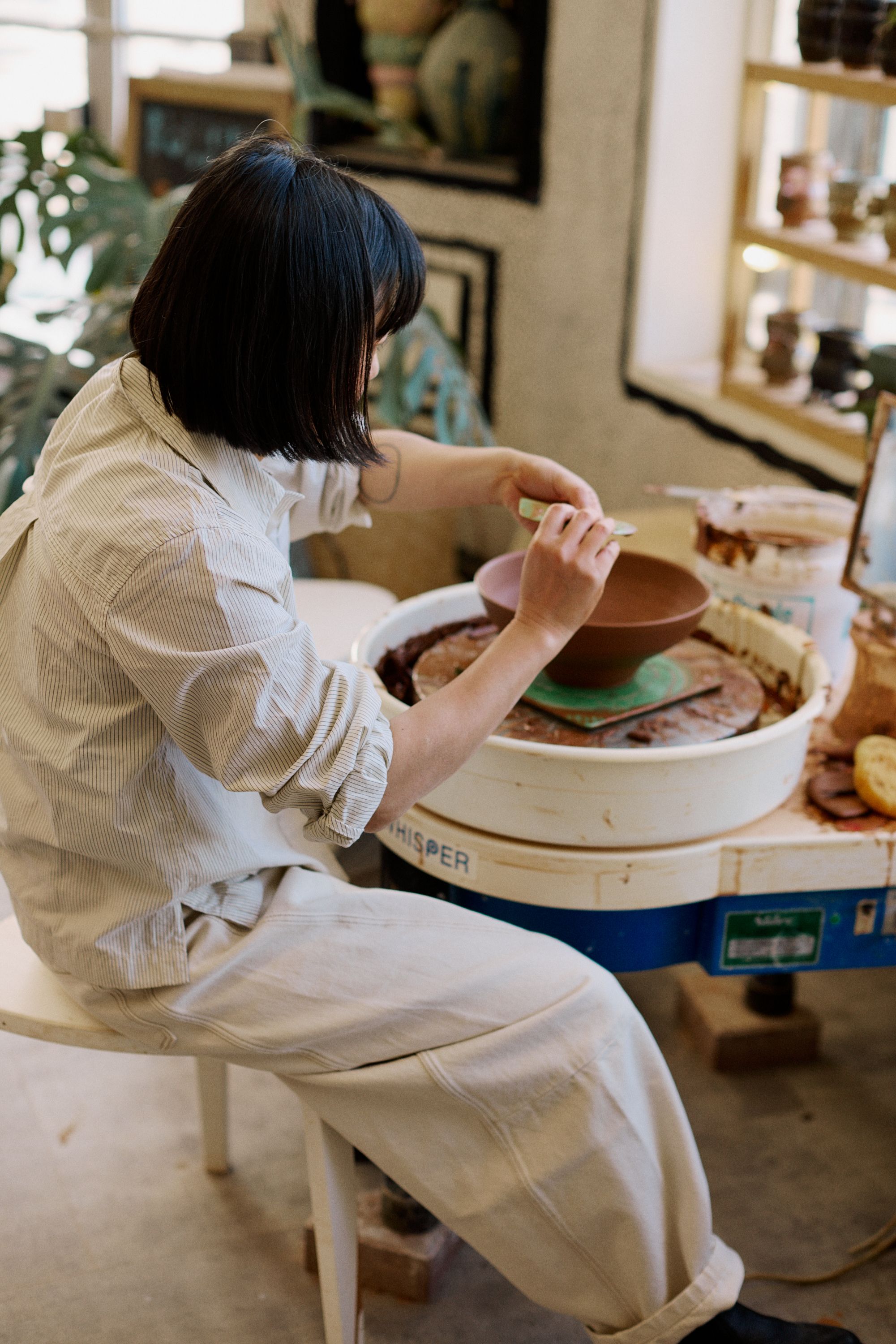 A woman throwing ceramic at a wheel and two vases, one in black earthware, one in golden resin, sitting next to three home accessories in red, gold, and black, against a beige background.