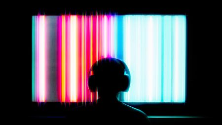 behind-the-head shot of a man watching TV with headphones on. a series of trippy vertical colored stripes are on the screen he watches