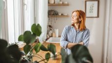 Woman with eyes closed looking towards sunlight coming through window in plant-filled kitchen