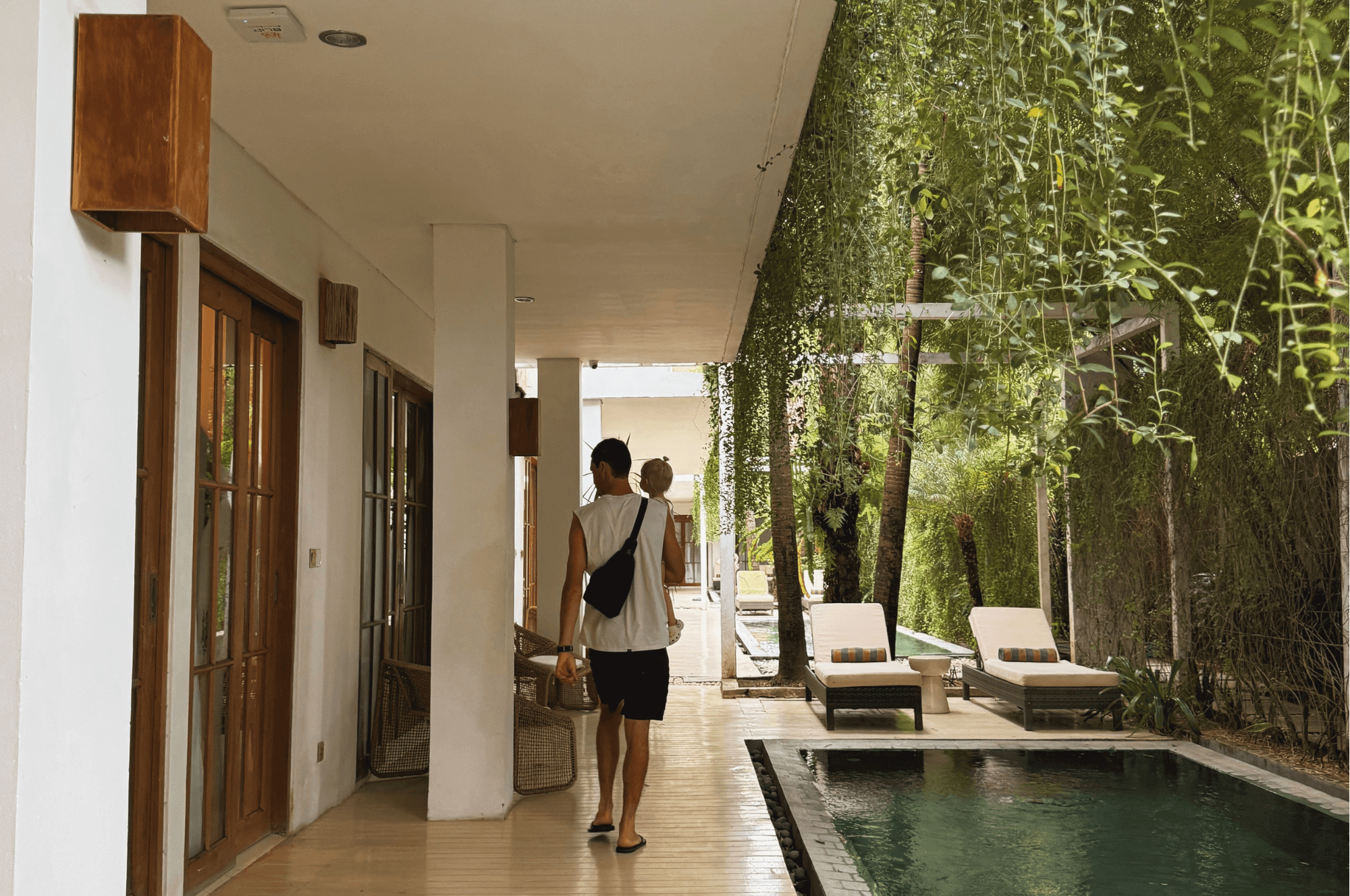 A person walks down a covered outdoor walkway next to a swimming pool, with lush green plants hanging overhead and lounge chairs visible by the water.
