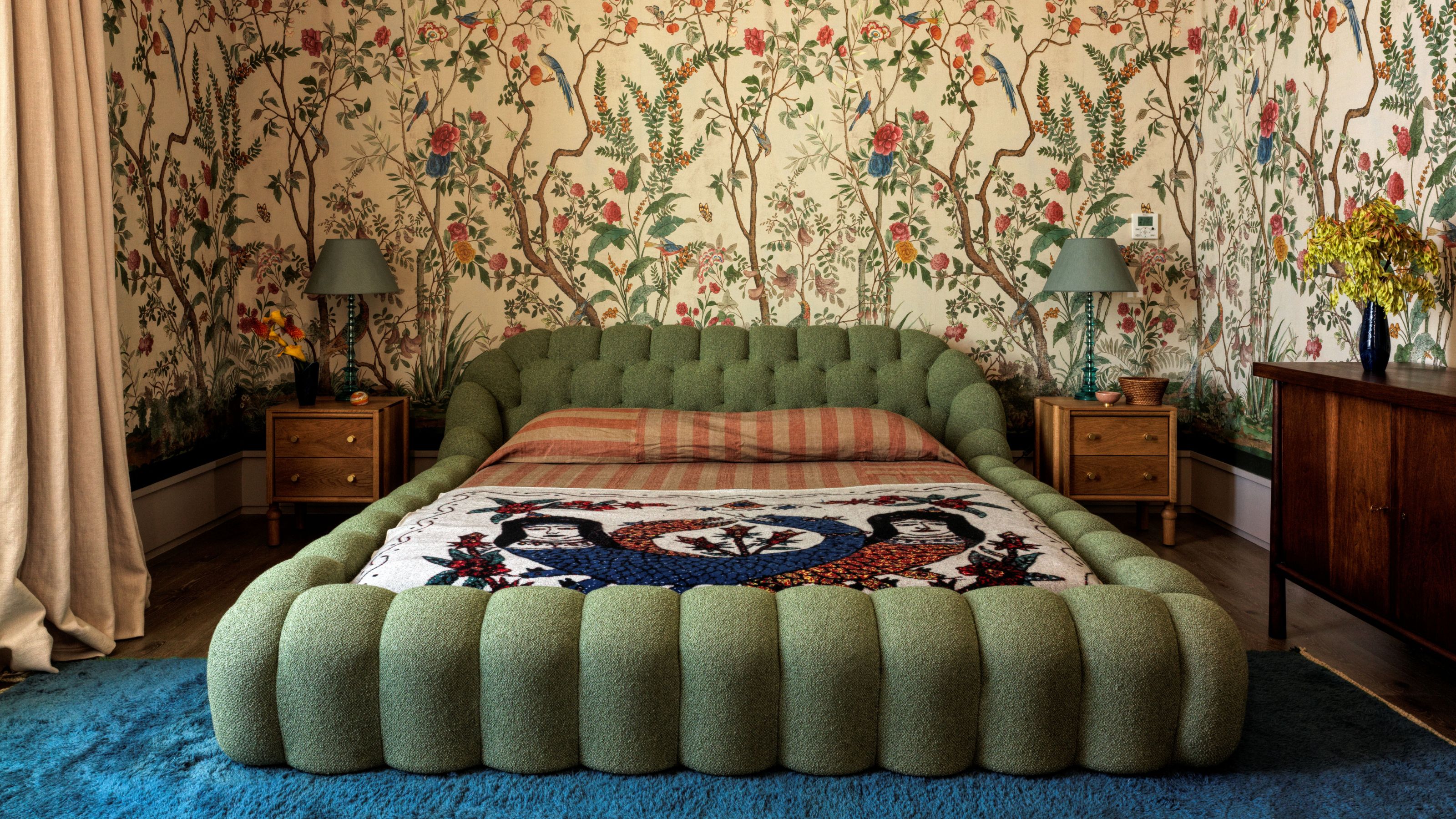 Bedroom with floral mural wallpaper on all walls and a green unholstered bed in the middle with two wooden side tables with green lamps