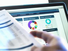 Person's hand is seen holding stocks and shares ISA document while looking at investment portfolio on laptop screen.