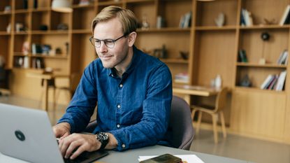 A man smiles as he works on his laptop in his large office.