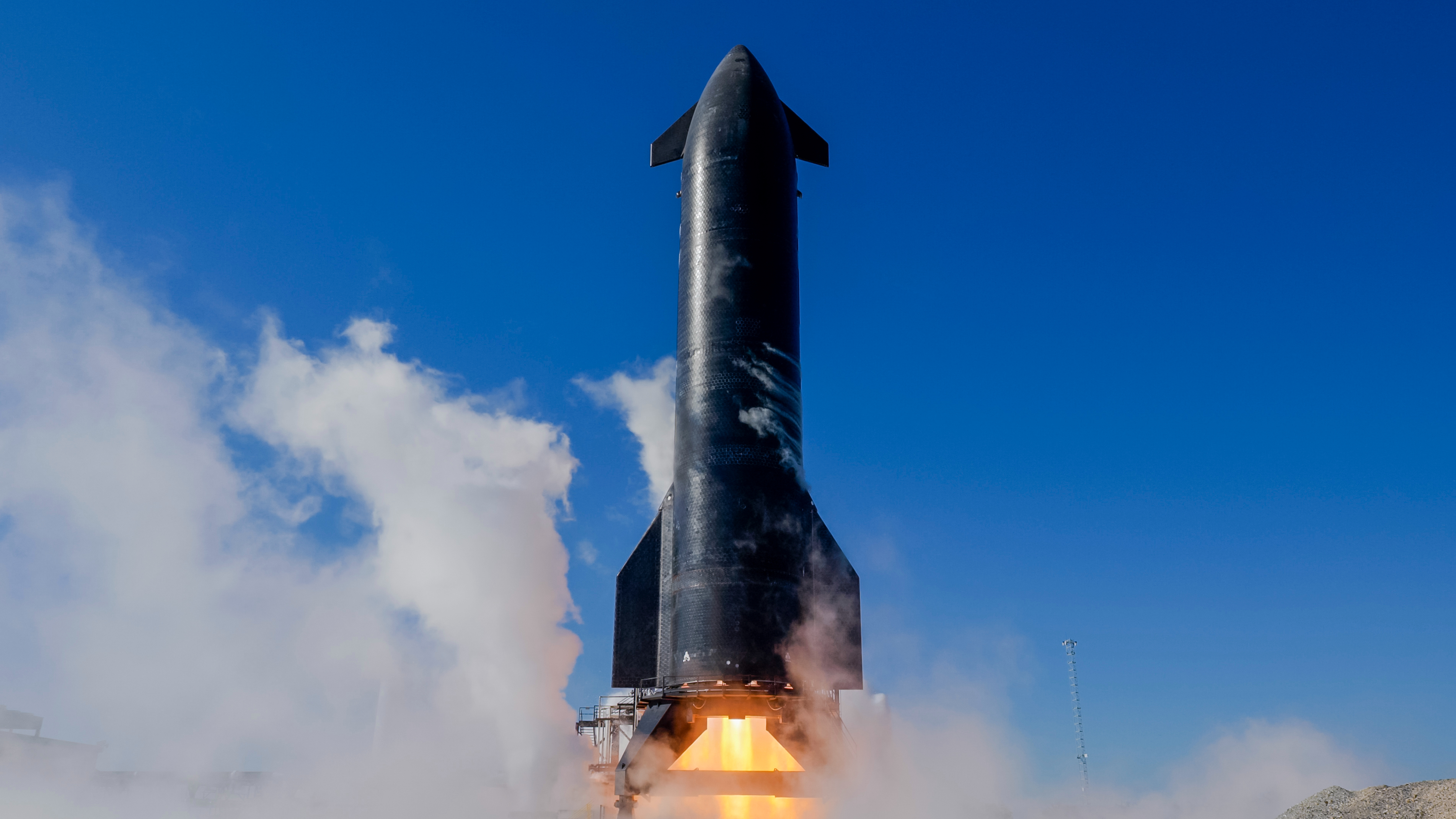 a tall silver spacecraft fires its engines on a test stand beneath a blue sky