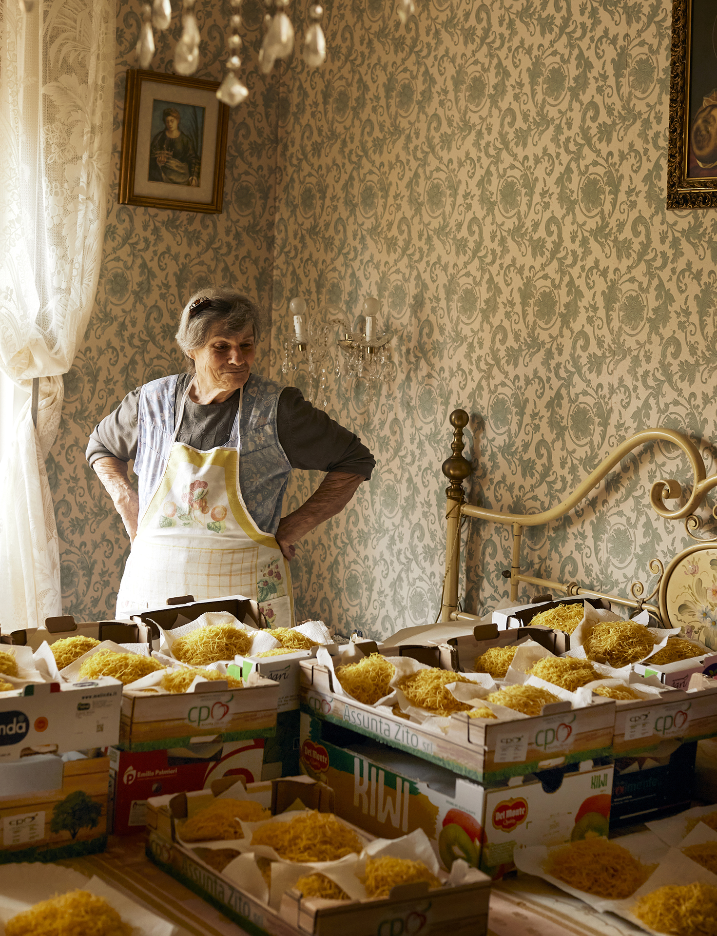 A woman stands in a vintage room filled with boxes of yellow, shredded pasta, illuminated by natural light from a nearby window