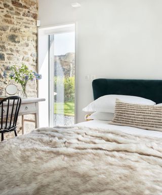 A bed with a beige blanket against white walls and exposed stonework.