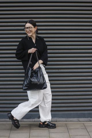 Maria Barteczko, wearing a black knitted polo shirt by Gitta Banko, white nylon cargo pants by Bershka, black gurkha fisherman sandals by Asos, a black hobo leather shopper by Chanel22, silver long crystal earrings by Chanel and black oversized Aviator glasses by Victoria Beckham, during a street style shooting at on May 17, 2023.