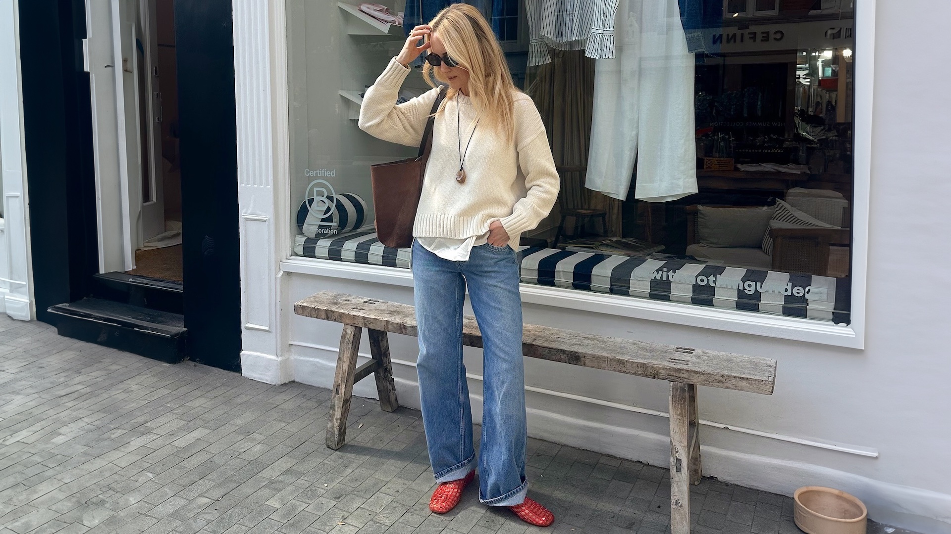 British style influencer Chloe Butler stands outside a shop in London wearing a cream sweater, pendant cord necklace, brown suede tote bag, cuffed jeans, and red jelly flats