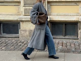 Danish art director Clara Dyrhauge walking on a cobblestone sidewalk wearing an oversize gray belted coat, a brown suede bag, straight-leg jeans, and black kitten-heel boots