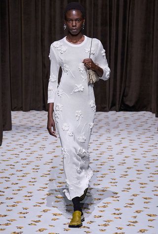 A snakeskin purse is shown in a photo of a model walking down Jil Sander's spring runway shows carrying a python shoulder bag styled with silver earrings, a white knit dress, and yellow loafers